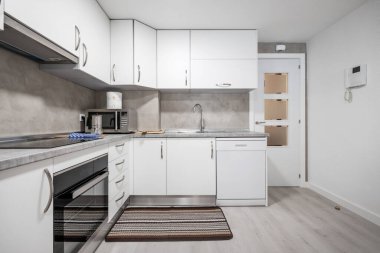 Aletleri olan büyük bir mutfak, gri tezgahlar ve ahşap döşemeli beyaz dolaplar, lavabonun yanındaki halı ve beyaz ahşap değirmen.