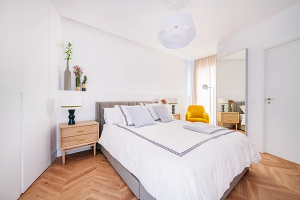 Açık renkli yastıklı çift kişilik yatak odası, açık ahşap komodinleri, çekmecelerden ve vazolardan oluşan hasır sandık ve dekoratif çiçekli vazolar ve kiralık bir dairedeki duvarda duran ayna.