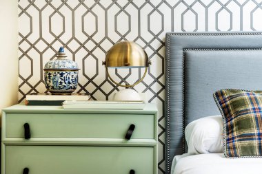 Bedroom with double bed with upholstered headboard with studs and gray fabric, pale green wooden bedside table, metal lampshade lamp and wallpapered walls