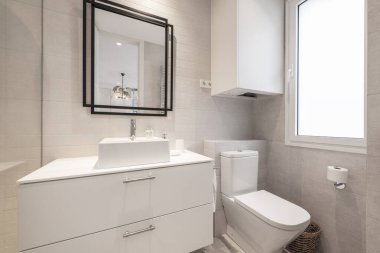 Parlak beyaz ahşap mobilyalı tuvalet, porselen lavabo ve siyah metal çerçeveli ayna.