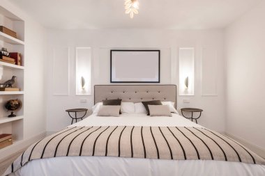 Yatak odası bej kumaşla döşenmiş, uyumlu battaniye, iki komodinli yatak odası ve alçı işçiliği rafı.