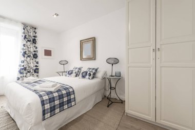 Çift kişilik yatak odası, rulo havlular, beyaz ahşap kapılı gardırop, yastıklar, battaniye ve uyumlu mavi perdeler ve lambalı ikiz komodinli yataklar.