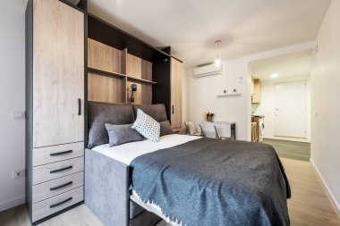 Rollaway bed inside sofa with cushions in short-term rental studio with wardrobe and light-wood floorboards