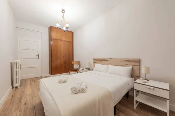 bedroom with double bed with rolled towels and built-in wardrobe with wooden doors and one-piece trunk, one-piece wooden headboard and twin white bedside tables