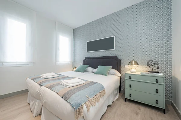 Çift yatak odalı soluk yeşil çekmeceler, uyumlu yastıklar, gri kumaşla döşenmiş yatak başlığı, duvarlarda süslemeli kağıt ve şeffaf beyaz panjurlu iki pencere.