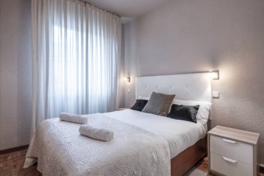 Beyaz deri kapitonlu yatak odası döşenmiş yatak başlığı, yastıklı çift kişilik yatak, rulo havlular ve ikiz komodinli masa.