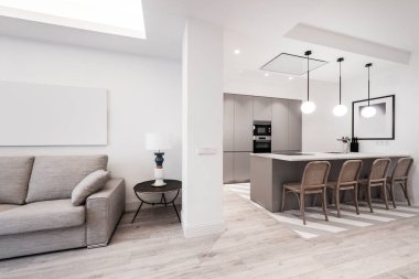 Beyaz taş tezgahlı mutfak, gri dolaplar, paslanmaz çelik aletler, eşleşen ada ve oturma odası gri kumaşla döşenmiş kanepe.