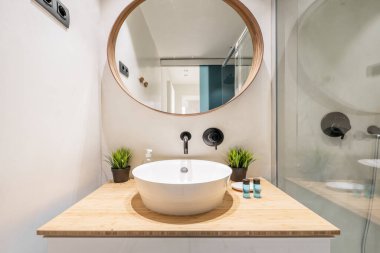 Parlak ahşap duvarlı banyo, parlak ahşap tezgah, dekoratif bitkiler ve ahşap çerçeveli dairesel ayna ve cam bölmeli duş kabini.