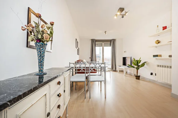 Square white dining table with sky blue metal chairs, wooden sideboard with black marble top, glass vase with dried flowers