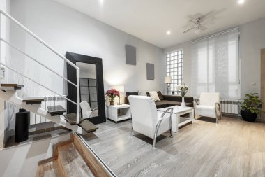Multi-storey loft and living room with brown fabric sofa with chaise longue, white side table, wooden flooring, paves wall, mirror on the wall and wooden steps with metal structure