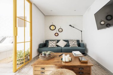Living room with vintage wooden side table with drawers, white brick wall, blue fabric 3 seater sofa with white cushions, wall mounted flat screen tv and yellow metal and glass doors