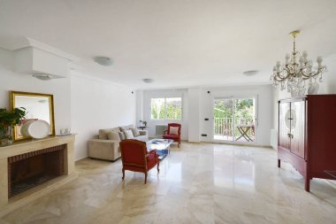 Şömineli oturma odası, mermer zemin, bej kanepe ve kırmızı koltuklar, ahşap gardırop ve bahçeli bir verandaya erişim.