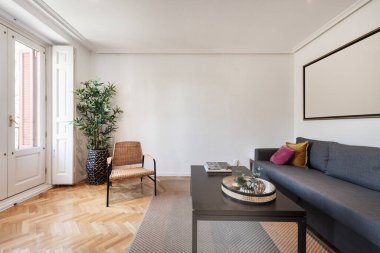 Kumaş kanepeli oturma odası, koyu ahşap masa, herringbone meşe parke, dekoratif bitki, hasır sandalye ve beyaz ahşap panjurlu balkonlar.