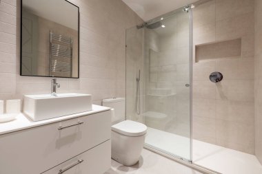Banyoda beyaz porselen lavabo, beyaz çekmeceler, siyah çerçeveli ayna ve cam kaygan duş kabini.