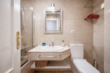 Duvarlarla uyumlu mermer tezgahlı banyo, gümüş çerçeveli ayna ve cam bölmeli duş kabini.