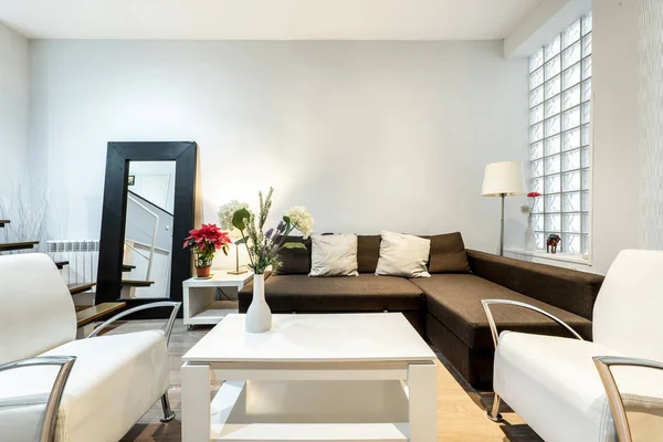 Living room with brown fabric sofa with chaise longue, white sideboard with flat screen tv, skylight wall with glass pavers, white wood side table and twin white leather armchairs