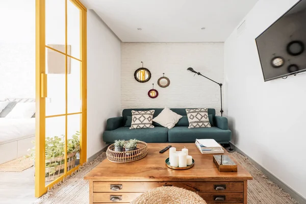 Living room with vintage wooden side table with drawers, white brick wall, blue fabric 3 seater sofa with white cushions, wall mounted flat screen tv and yellow metal and glass doors