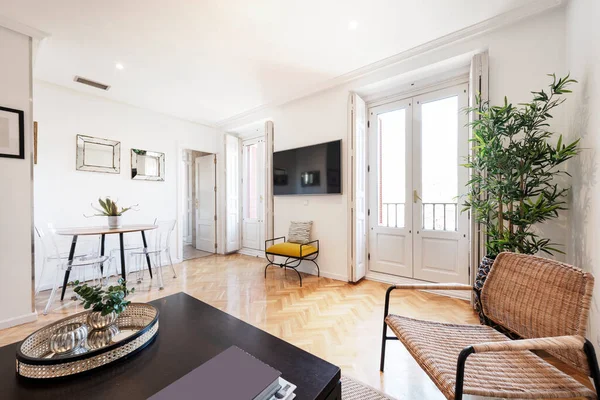 Yuvarlak ahşap yemek masası, koyu ahşap yan masası, ringa kemiği meşesi parke, dekoratif bitki, hasır koltuk ve beyaz ahşap panjurlu balkonları olan oturma odası.