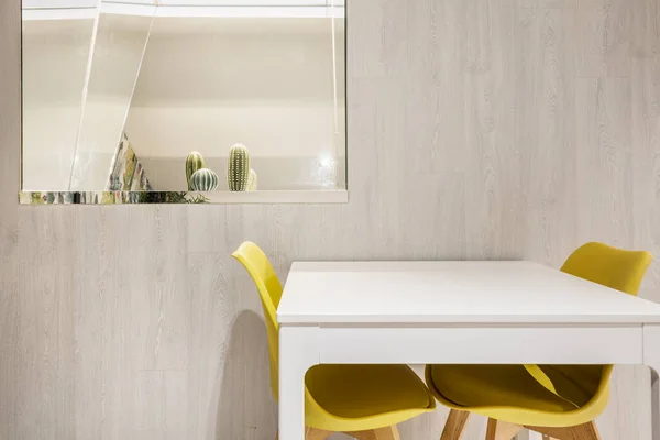 White square wooden dining table with yellow resin chairs and beaded framed mirror on the wall