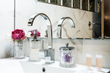 Beyaz porselen lavabosu olan bir vestiyerin köşesinde krom musluğu ve cam aksesuarları olan eğimli bir aynanın altında.