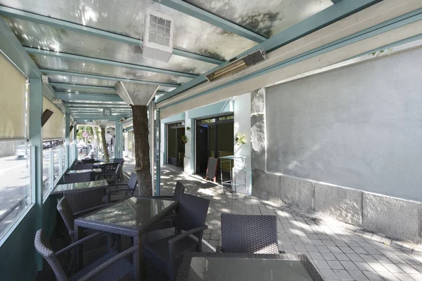 Porch of a light blue restaurant on the sidewalk of a street with rattan tables and chairs