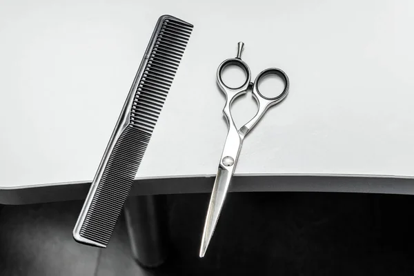 Stainless steel scissors and black barber\'s comb on a white wooden countertop