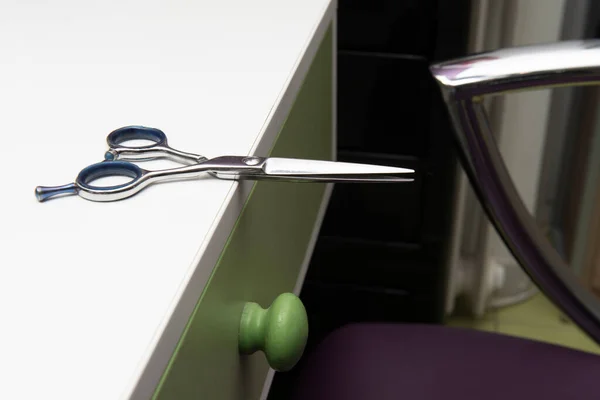 Stainless steel scissors and a black barber comb on a large white wooden counter with a green drawer and a chair with metal arms