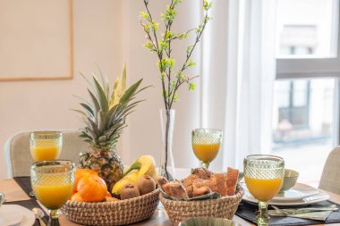 Round wooden dining table with breakfast service with sweets, orange juice and assorted fresh fruits and a decorative vase with a plant