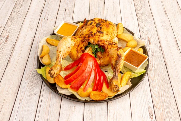 Charcoal grilled chicken with potato wedges, sauces, lettuce, parsley, lemon wedges and fresh red pepper on a black porcelain tray