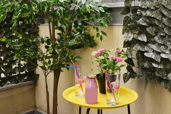 Corner of a terrace with artificial decorative plants, yellow metal side table with glasses and strawberry juice