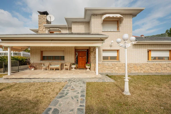 Facade of a single-family home with a covered porch with wicker furniture and gardens with stone paths