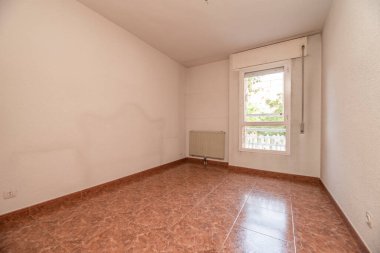 Kırmızımsı taş döşemeli boş oturma odası, duvarda beyaz radyatör ve manzaralı pencere.