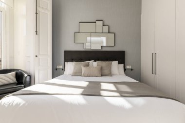 Yastık ve yastıklı büyük boy yatak odası, koyu kahverengi kumaşla döşenmiş yatak odası, beyaz kapılı gardırop ve beyaz ahşap kapılı balkon ve panjurlar.