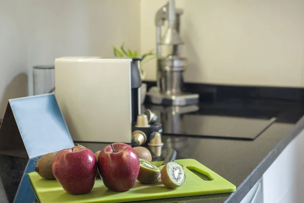 Corner of a kitchen with a black stone countertop with a white capsule coffee machine, designer juicer and a green Teflon board with sliced red apples and kiwis