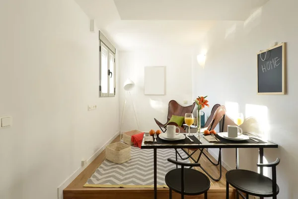 Small room with table for breakfast, orange juice and fruit, leather armchairs, matching black metal chairs and white aluminum window