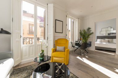Halılı bir evin oturma odası, çiçekli bir yan masası olan gri kumaştan bir aşk koltuğu, pencereli birkaç balkon, beyaz ahşap panjurlar ve parlak sarı kumaştan bir kanat sandalyesi.