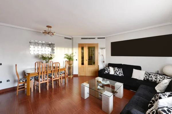 Living room of a residential home with a wooden dining table with matching chairs, a corner sofa covering two walls upholstered in black fabric and reddish stoneware floors