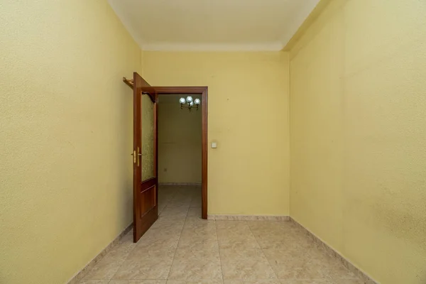 Empty small room is square tile stoneware floor, yellow painted wall with gotelet and reddish wooden door with glass window