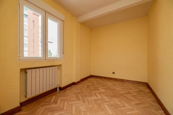 Empty room with aluminum window, radiator in a niche, varnished oak parquet and soft yellow painted walls