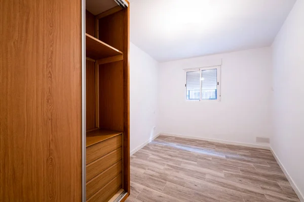 Renovated room with built-in wardrobe with sliding doors in cherry wood color, interior chest of drawers, white aluminum window and aluminum radiator on the wall