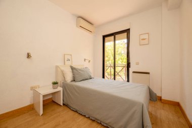 Gri yastıklı bir yatak odası, aynı yatak örtüsü, ahşap döşemeler ve bahçeye bakan bir balkon.