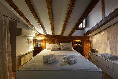 Çift kişilik yatak odası, eğimli tavanlar, açıkta duran ahşap kirişler, aynı yastıklar, aynı yatak örtüsü ve ahşap pencere.