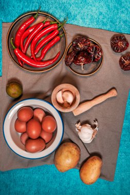Bir tabak çiğ yumurta, çiğ patates, ahşap havanda sarımsak, kurutulmuş biber ve gri kumaş ve mavi arka planda kırmızı biberle dolu bir hayat.