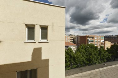 Açık renkli duvarı, küçük beyaz alüminyum pencereleri ve yağmurlu bulutları olan bir evin cephesi