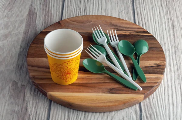 Sarı kullan-at kağıt bardaklar, beyaz plastik çatallar, yuvarlak ahşap bir stantta yeşil kaşıklar ve gri bir arka plan. Yüksek kalite fotoğraf
