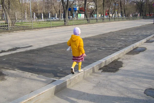Sarı ceketli küçük bir kız kaldırım kenarındaki kaldırımda yürüyor.