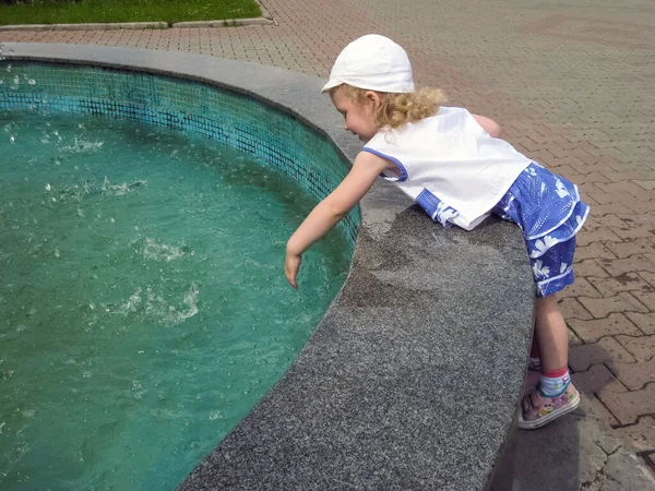 Mavi beyaz elbiseli küçük bir kız ve beyaz şapkalı bir kız şehir parkındaki çeşmenin suyuna uzanıyor. Yaz. Gün.