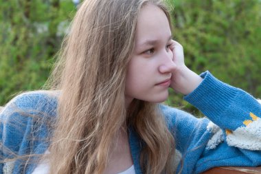 Mavi hırkalı, gevşek sarı saçlı genç bir kız yeşil bulanık bir arka planda eli tarafından desteklenen mesafeye düşünceli bir şekilde bakar. Yaz. Yakın plan.