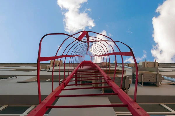 Bulutlu mavi gökyüzüne karşı gri bir binanın ön cephesinde kırmızı bir yangın merdiveni. Bakış açısı. Düşük görünüm açısı