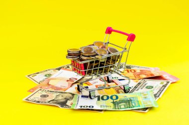 İçinde bozuk paralar, kâğıt para ve sarı arka planda dolar banknotları olan mini alışveriş sepeti. Finans ve tasarruf kavramı.